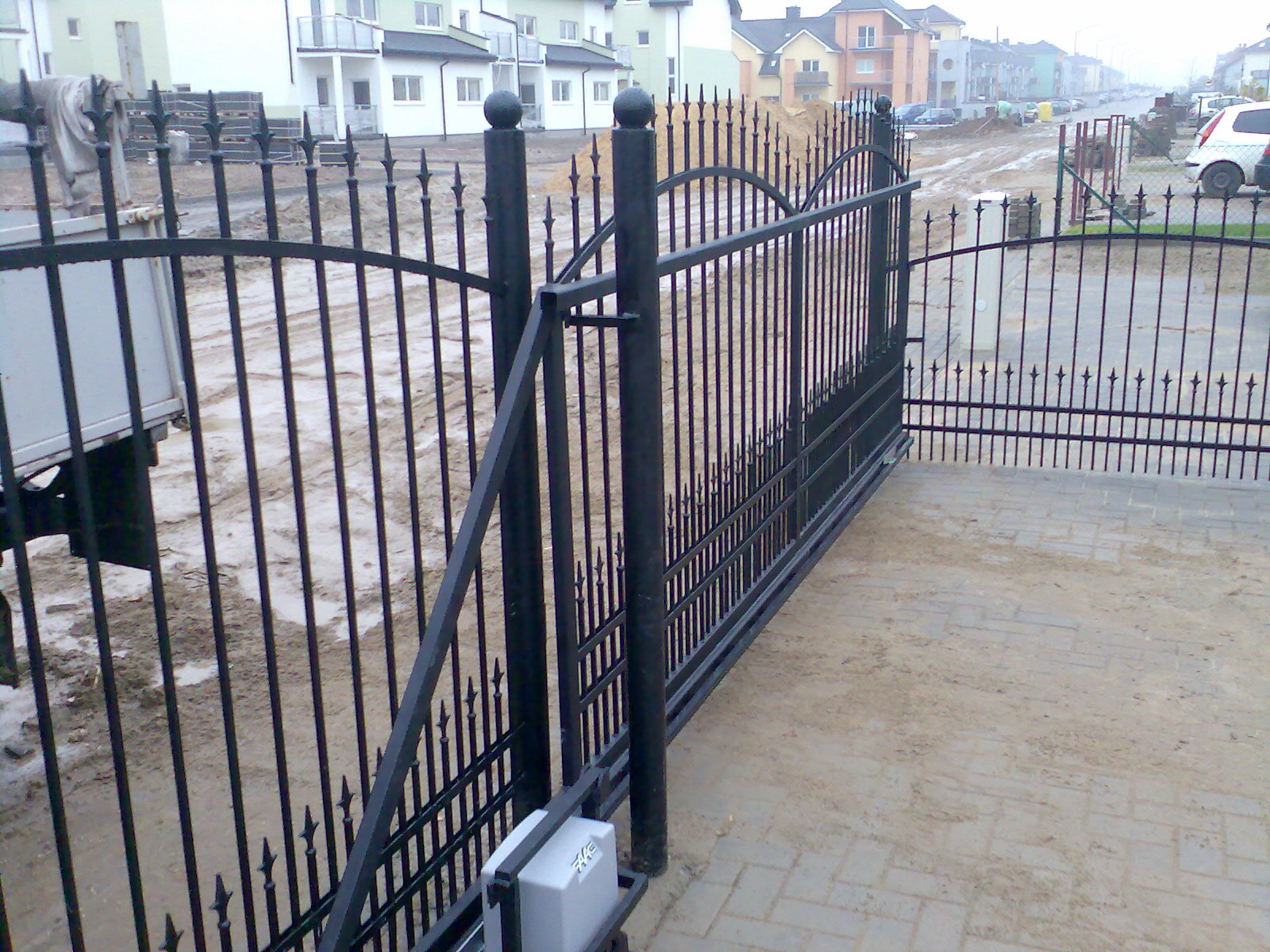 Czarna, metalowa brama wjazdowa przesuwna z automatyką, widoczny mechanizm otwierający, na tle nowo wybudowanych domów i błotnistej drogi.